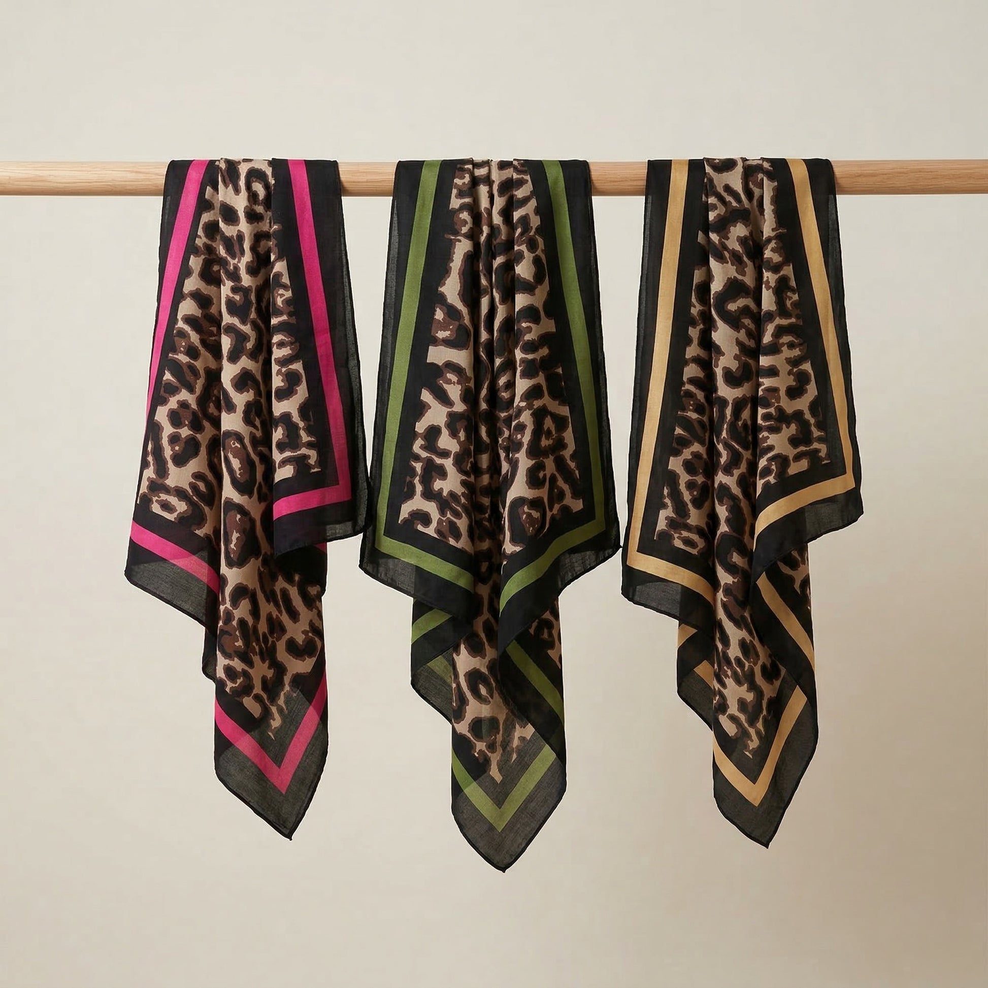Three leopard print scarves with color-blocked borders hanging on a rod against a beige background.